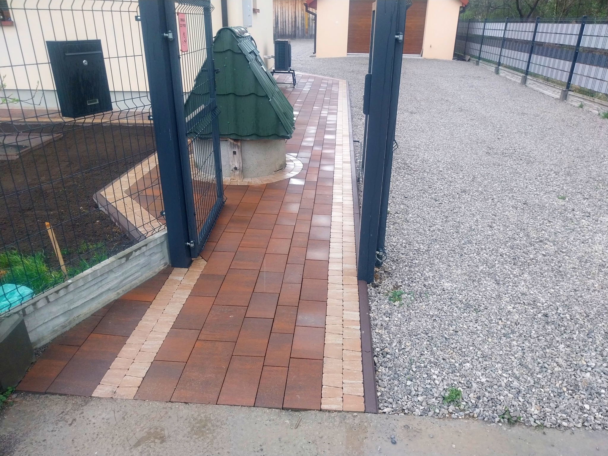 Ścieżka z brązowej kostki brukowej z beżowymi akcentami, prowadząca od furtki do studni z zielonym daszkiem, obok wysypany żwir.