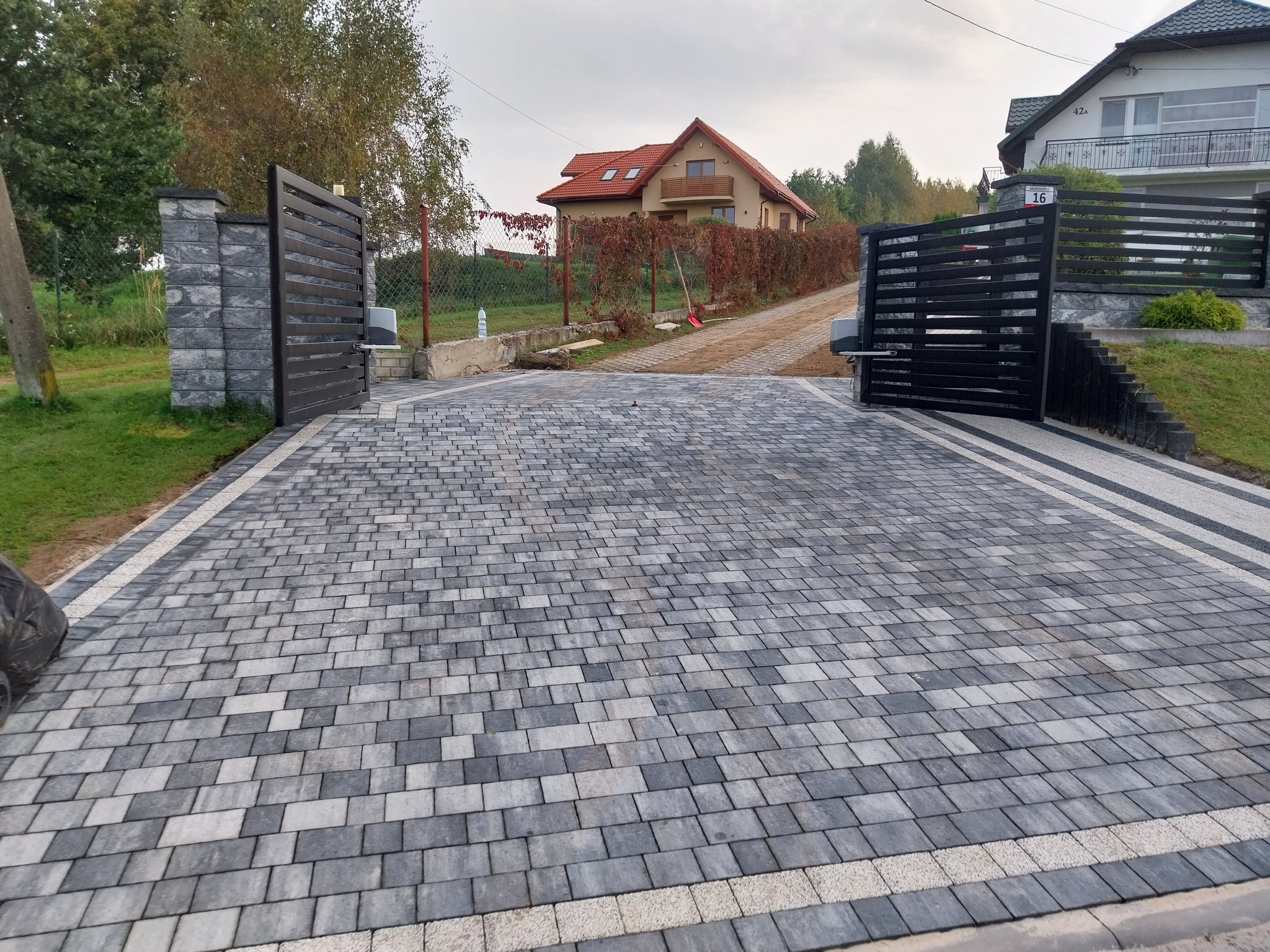 Ułożona kostka brukowa w odcieniach szarości z jasnymi obrzeżami, tworząca podjazd do posesji z automatyczną bramą w tle.