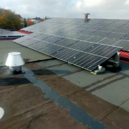 Panel słoneczny zamontowany na płaskim dachu z widocznymi elementami wentylacyjnymi i uszczelnieniami, w tle budynki mieszkalne i drzewa.