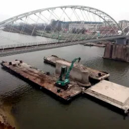 Demontaż filaru mostu z użyciem barki i koparki na rzece, w tle widoczny nowy most łukowy i budynki miejskie.