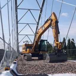 Żółta koparka CAT na żwirowym podłożu z białym materiałem geowłókniny, widoczna stalowa konstrukcja mostu w tle.