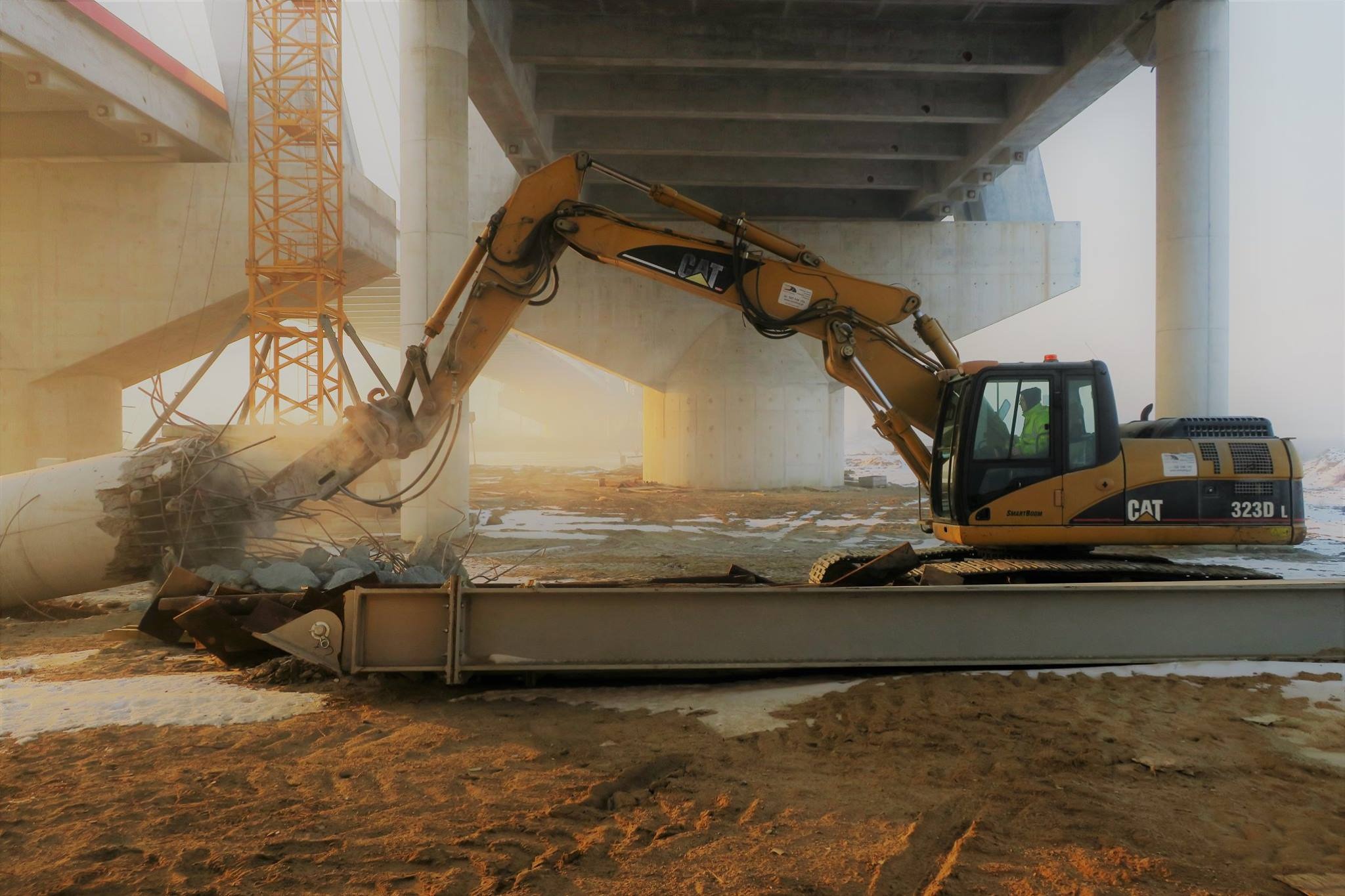 Żółta koparka CAT 323D L kruszy betonowy filar pod wiaduktem w pochmurny dzień, widoczny żuraw i resztki śniegu na ziemi.