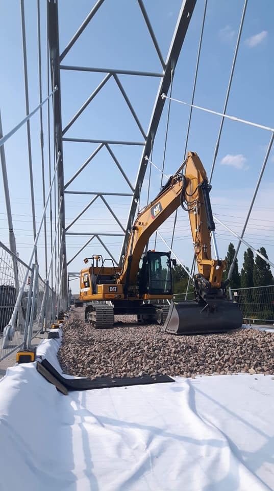Żółta koparka CAT na żwirowym podłożu z białym materiałem geowłókniny, widoczna stalowa konstrukcja mostu w tle.