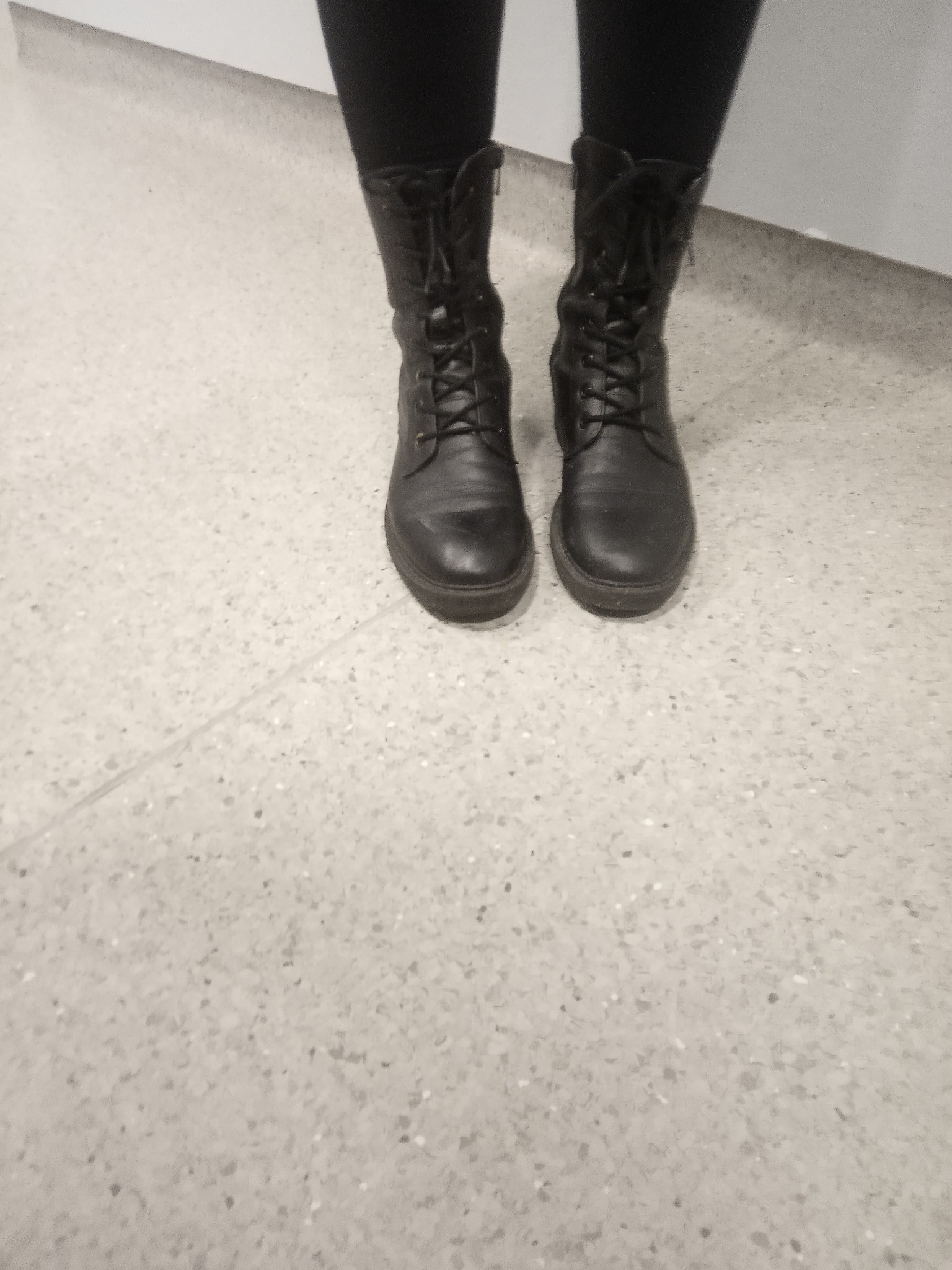 Osoba w czarnych legginsach i sznurowanych, skórzanych botkach stoi na jasnej, nakrapianej podłodze.