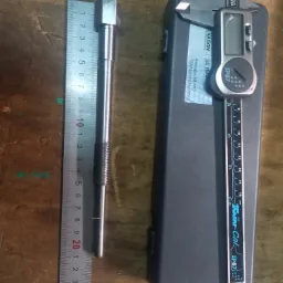 Metalowy trzpień z gwintowanym fragmentem, zmierzony linijką i suwmiarką cyfrową na tle drewnianego stołu warsztatowego.