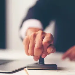 Dłoń przyciska stempel na dokumencie, widoczny fragment laptopa w tle.