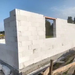 Wznoszenie ścian zewnętrznych domu z białych bloczków na fundamencie, widoczne otwory okienne z drewnianym nadprożem, w tle zieleń drzew i błękitne niebo.