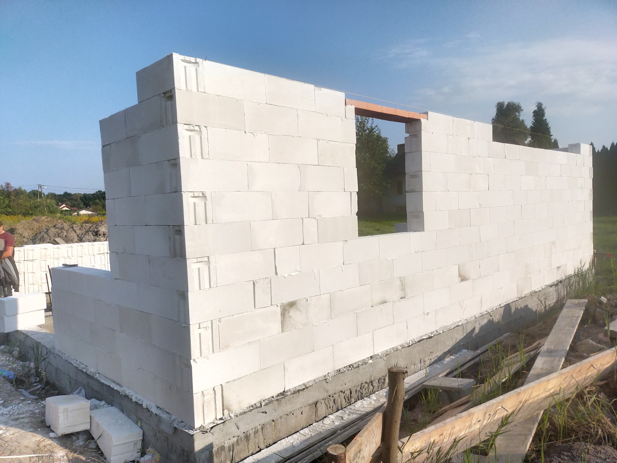 Wznoszenie ścian zewnętrznych domu z białych bloczków na fundamencie, widoczne otwory okienne z drewnianym nadprożem, w tle zieleń drzew i błękitne niebo.