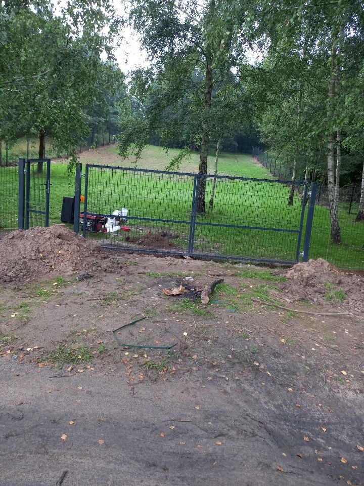 Szara metalowa brama i ogrodzenie panelowe w trakcie montażu, widoczne narzędzia i kopiec ziemi, w tle zielony trawnik i drzewa.