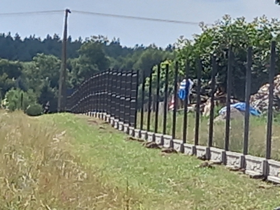 Długi, ciemny, panelowy płot z betonową podmurówką, otoczony trawą i drzewami w tle, z niebieskimi plandekami widocznymi za ogrodzeniem.
