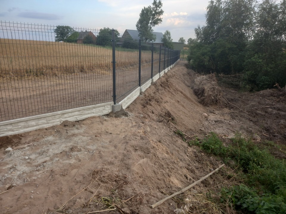 Długi, panelowy płot z podmurówką o fakturze kamienia, oddzielający pole od terenu z wykopem ziemnym i roślinnością.