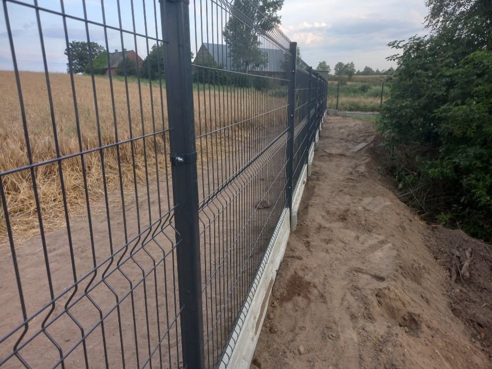 Panelowe ogrodzenie z podmurówką betonową na skraju pola zboża i zarośli, widok z perspektywy ziemi.