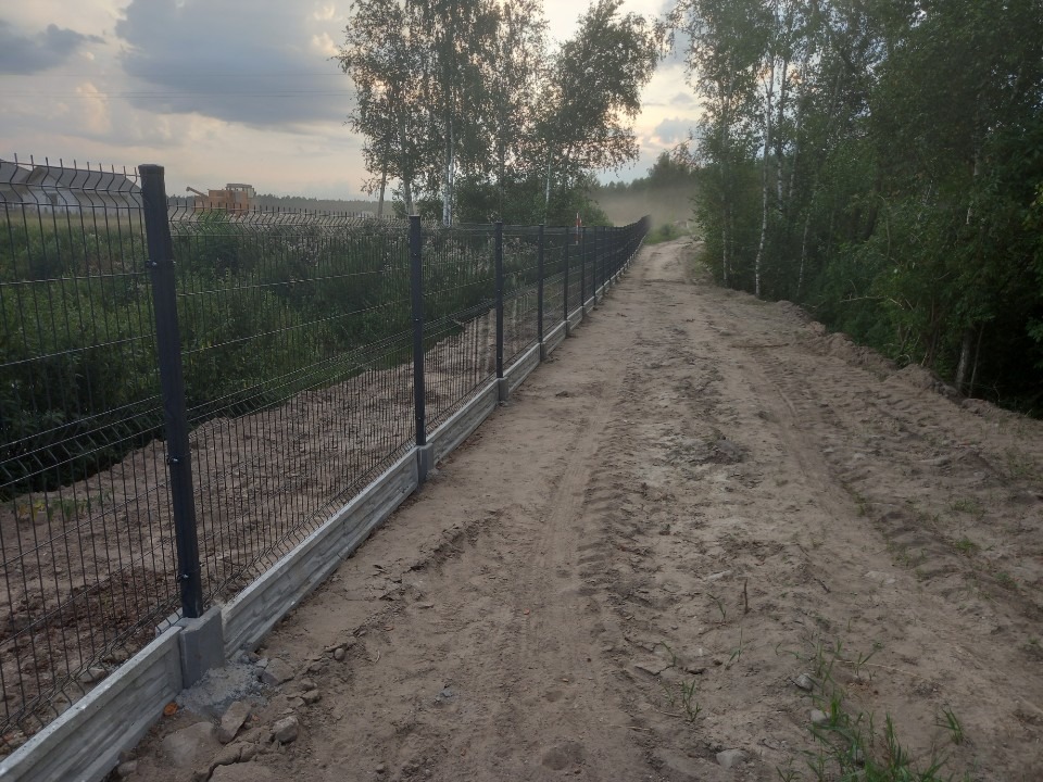 Długa, metalowa siatka ogrodzeniowa z betonową podmurówką, biegnąca wzdłuż polnej drogi z widocznymi śladami opon, z drzewami w tle i koparką w oddali.