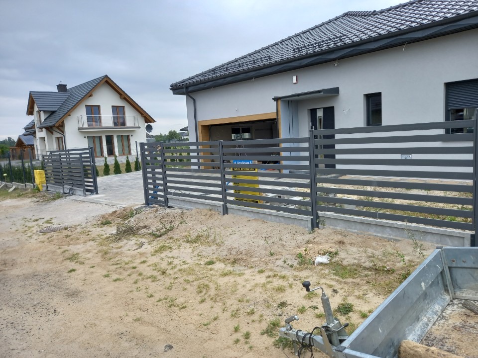 Nowoczesne, poziome ogrodzenie panelowe w kolorze grafitowym, z bramą przesuwną, przed nowym domem z szarą elewacją i ciemną dachówką, widoczny fragment przyczepy.