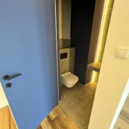 Nowoczesna toaleta w Kościerzynie z niebieskimi drzwiami, płytkami imitującymi lastryko, podświetleniem LED i drewnianą podłogą na zewnątrz.