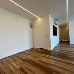 Nowoczesne wnętrze z drewnianą podłogą, białymi ścianami i minimalistycznym oświetleniem LED w suficie, klimatyzacja i czarna abstrakcyjna grafika na ścianie.