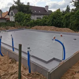 Przygotowany fundament pod budynek z widoczną izolacją termiczną i wystającymi rurami instalacyjnymi, otoczony piaskiem i zarysami przyszłego budynku w tle.