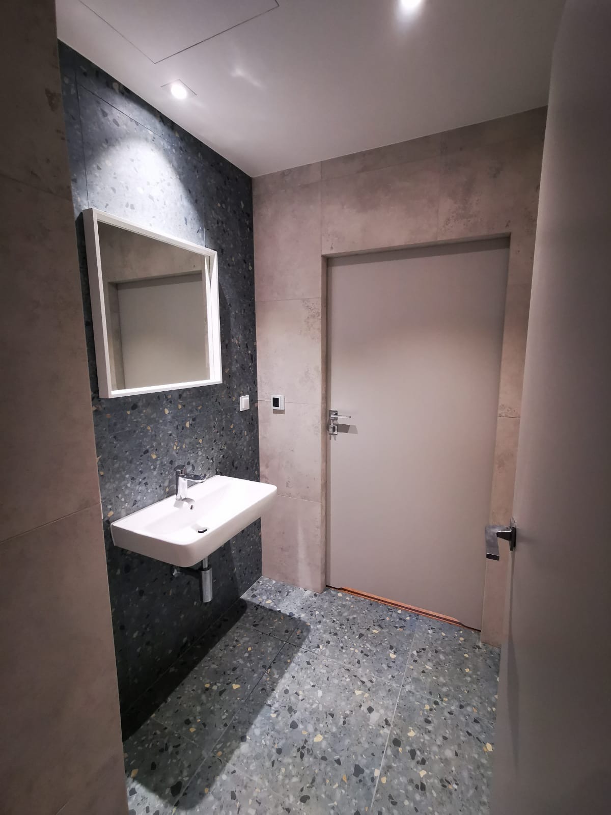 Wykończona łazienka z umywalką nablatową, lustrem w białej ramie i płytkami imitującymi lastryko na ścianach i podłodze. Drzwi wewnętrzne w kolorze szarym.