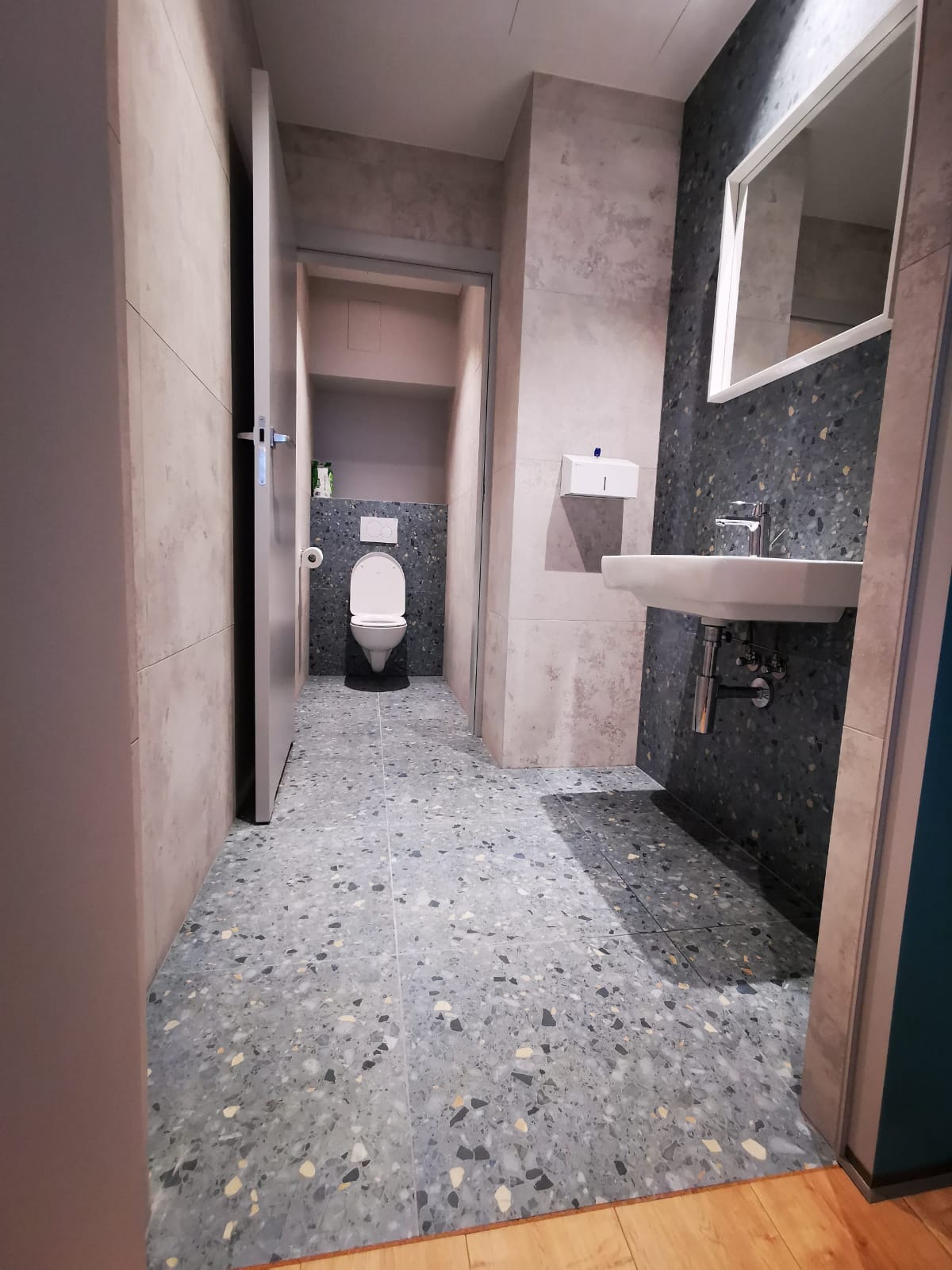 Wykończona łazienka z toaletą i umywalką, podłoga i ściany wyłożone płytkami imitującymi lastryko w odcieniach szarości i błękitu, widoczny fragment drewnianej podłogi przy wejściu.