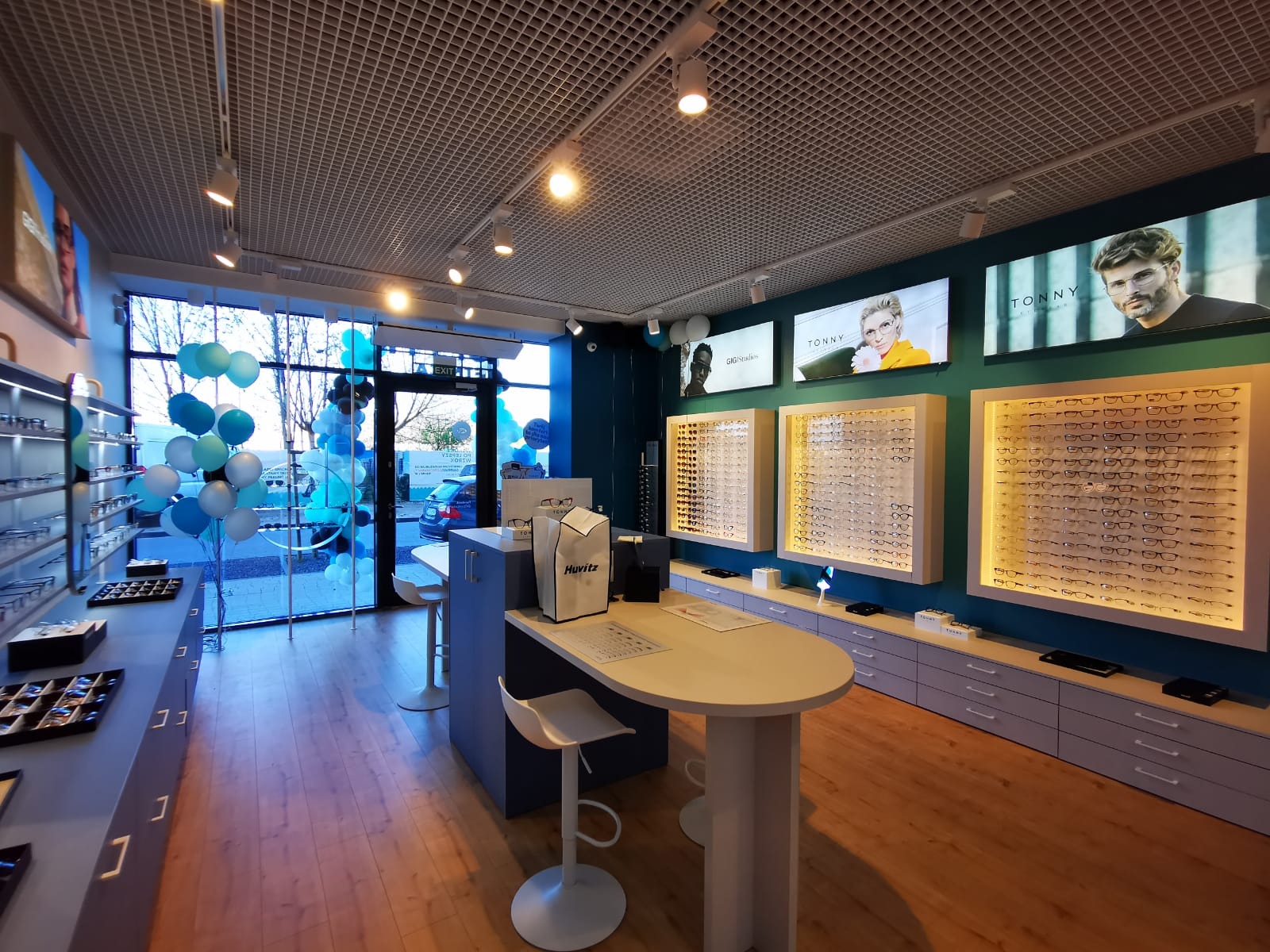 Wnętrze sklepu optycznego z ekspozycją okularów w ramkach na ścianach, stolikiem obsługi klienta i dekoracjami z balonów.