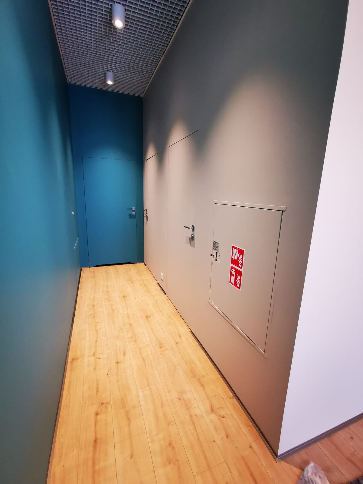 Korytarz biurowy z drewnianą podłogą, pomalowanymi ścianami w kolorze turkusowym i szarym, oraz ukrytymi drzwiami i szafką przeciwpożarową