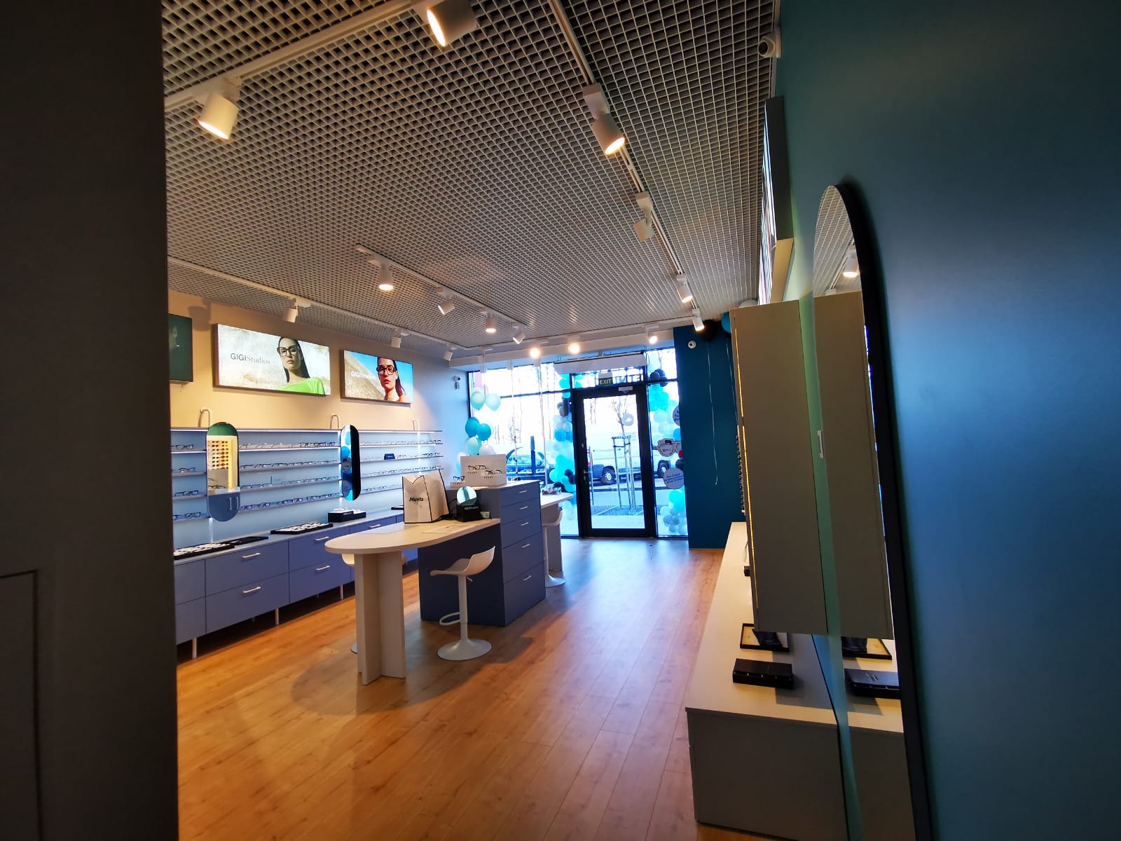 Wnętrze nowoczesnego salonu optycznego, z ekspozycją oprawek na niebieskich półkach, białymi stolikami i krzesłami, oraz reklamami na ścianach pod sufitem z kratką.