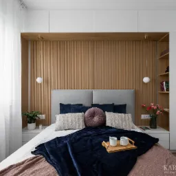 Nowoczesna sypialnia z drewnianą ścianą z lameli, szarą tapicerowaną ramą łóżka, dekoracyjnymi poduszkami, ciemnoniebieskim pledem, drewnianą tacą z dwoma kubkami oraz białymi szafkami w zabudowie.