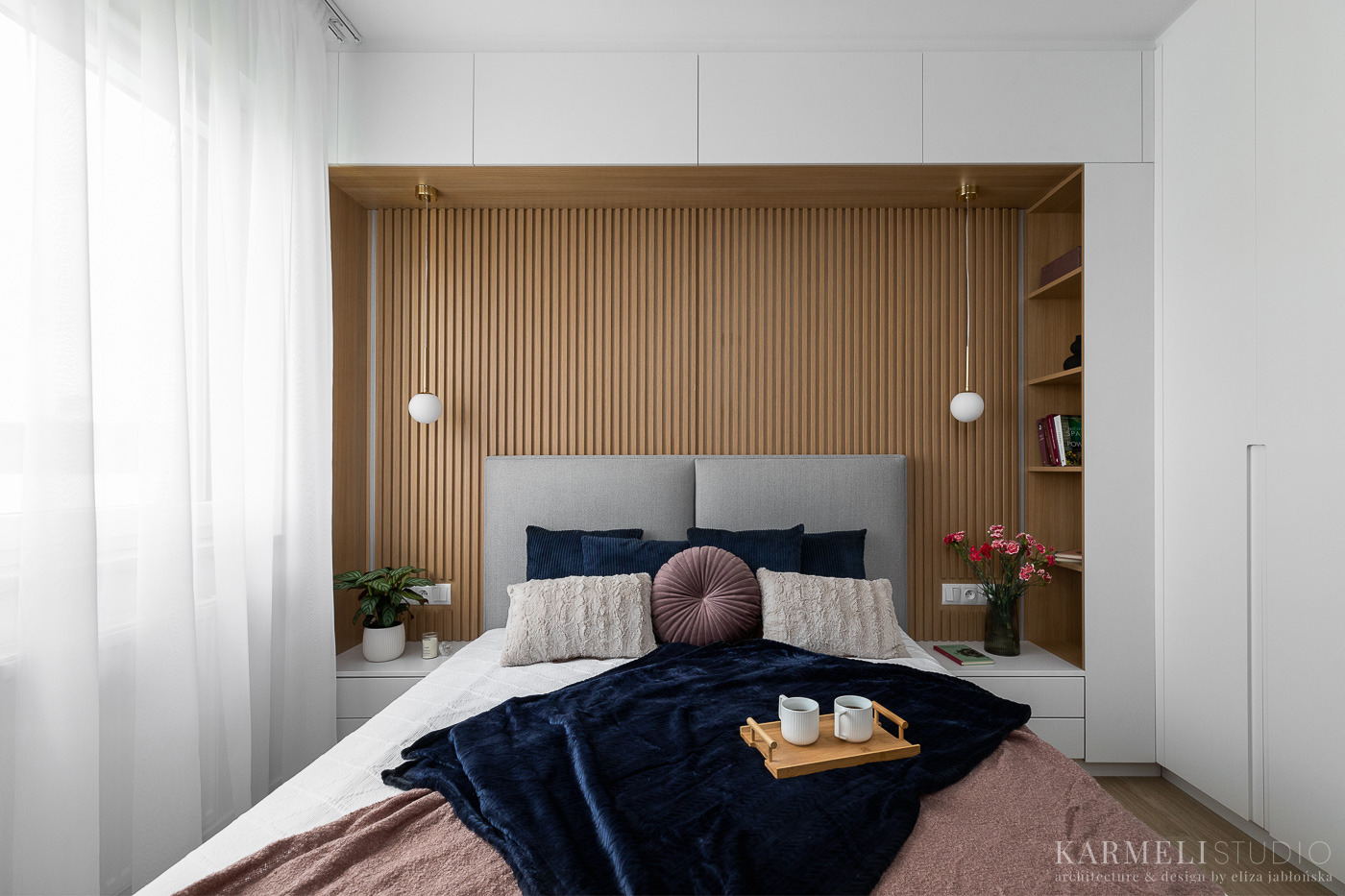 Nowoczesna sypialnia z drewnianą ścianą z lameli, szarą tapicerowaną ramą łóżka, dekoracyjnymi poduszkami, ciemnoniebieskim pledem, drewnianą tacą z dwoma kubkami oraz białymi szafkami w zabudowie.