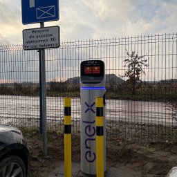 Metis Intech Sp. z o.o. - Stacja ładowania pojazdów elektrycznych Enel X z dwoma słupkami ochronnymi, umieszczona na parkingu z widocznym znakiem informacyjnym dla pojazdów elektrycznych.