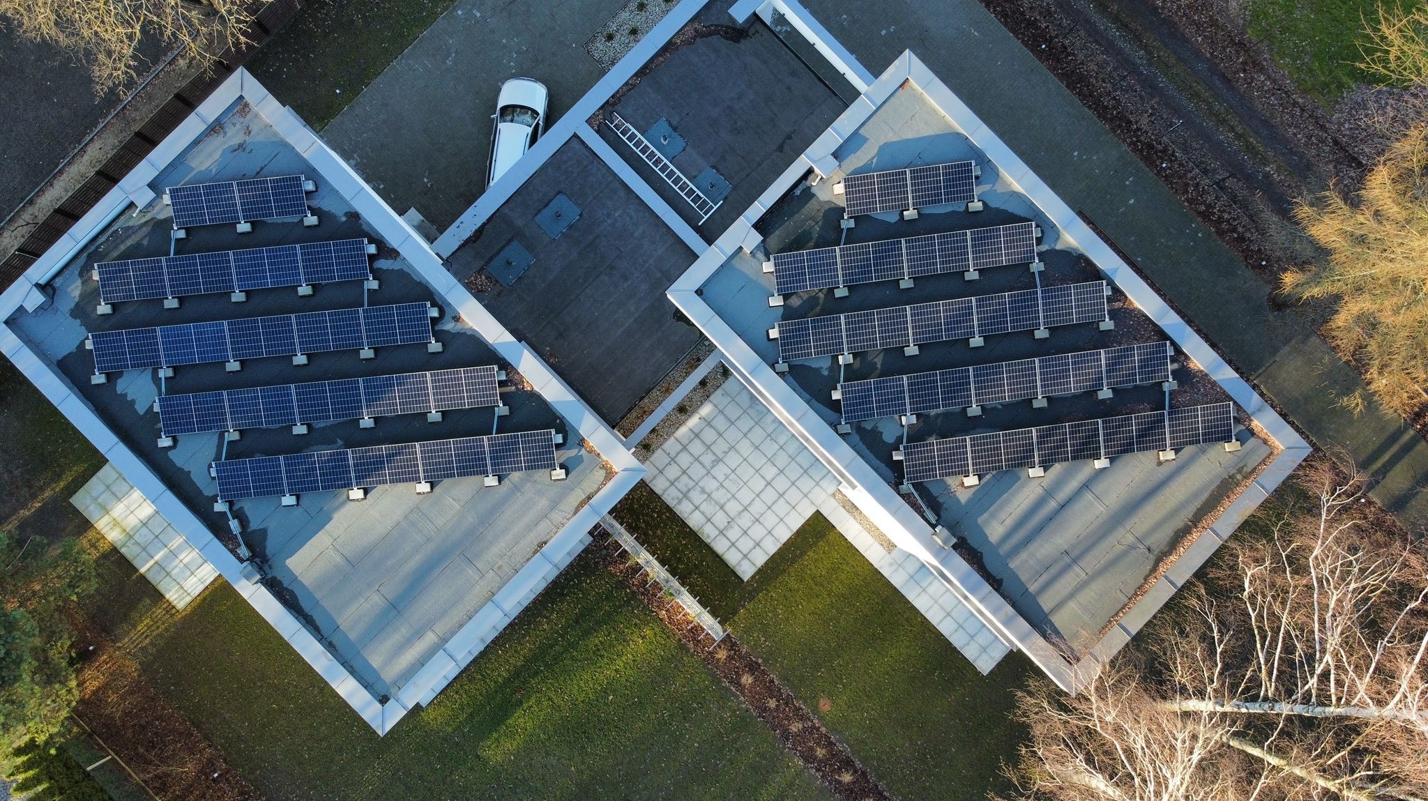 Widok z lotu ptaka na dach budynku z panelami słonecznymi ułożonymi w rzędach, obok zaparkowany samochód i drabina oparta o dach.