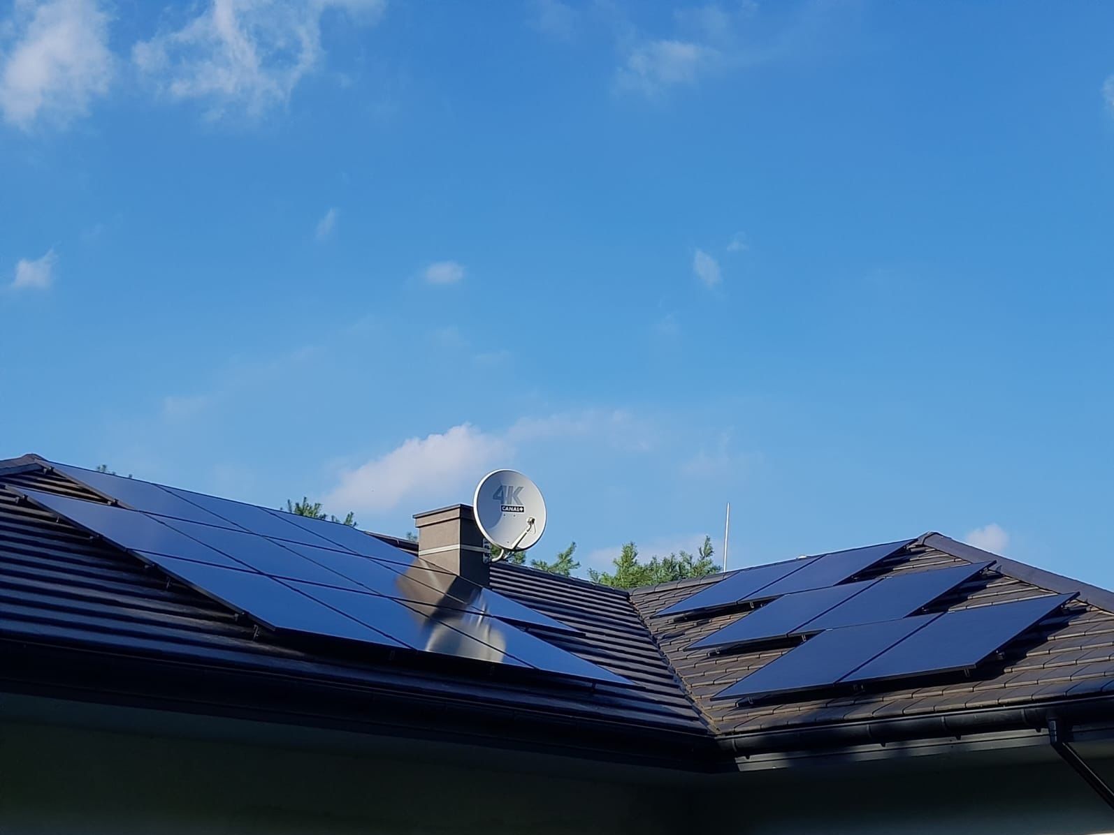 Panele słoneczne na brązowym dachu domu jednorodzinnego z anteną satelitarną na kominie, błękitne niebo w tle.