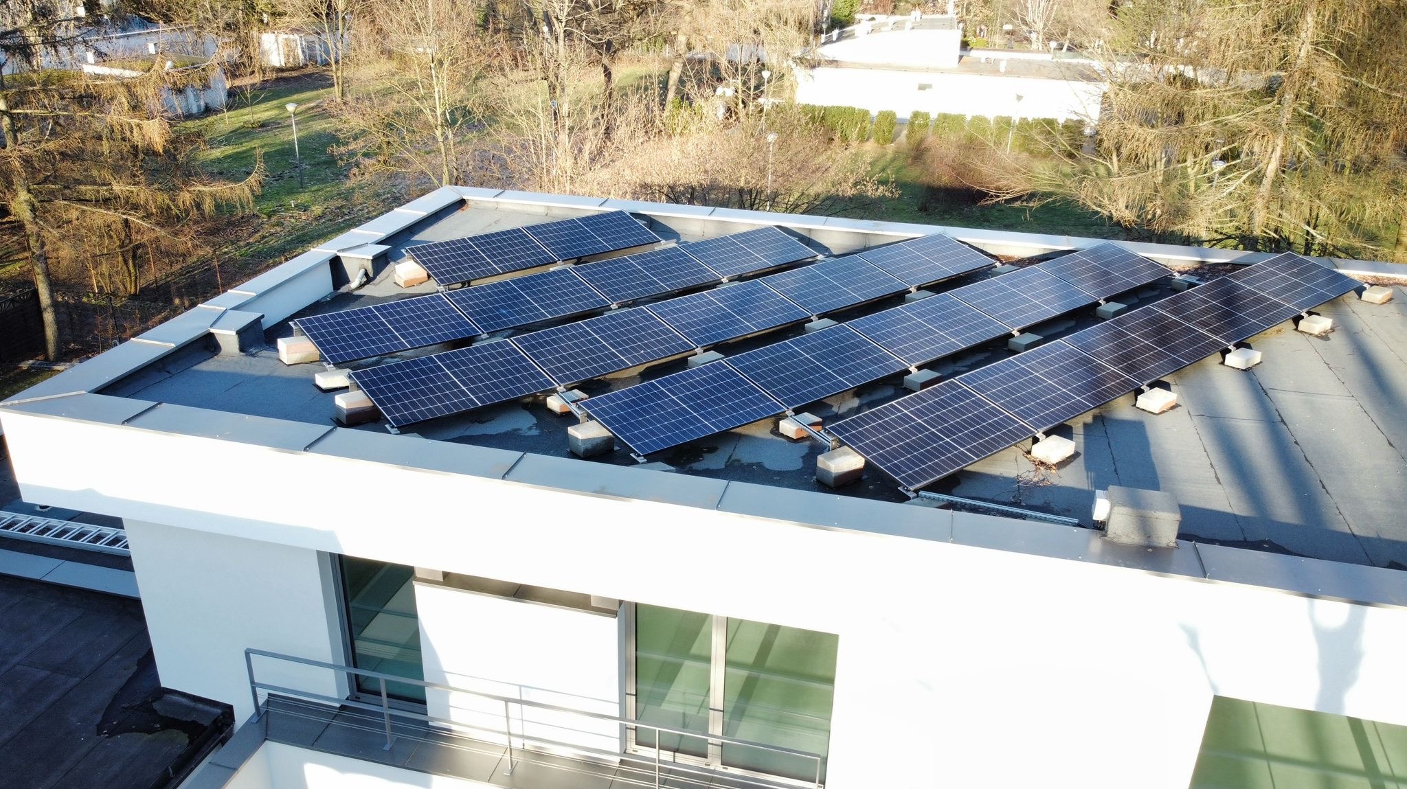 Rząd paneli słonecznych na płaskim dachu budynku, zamontowanych na betonowych wspornikach, z widocznym balkonem i otoczeniem drzew.