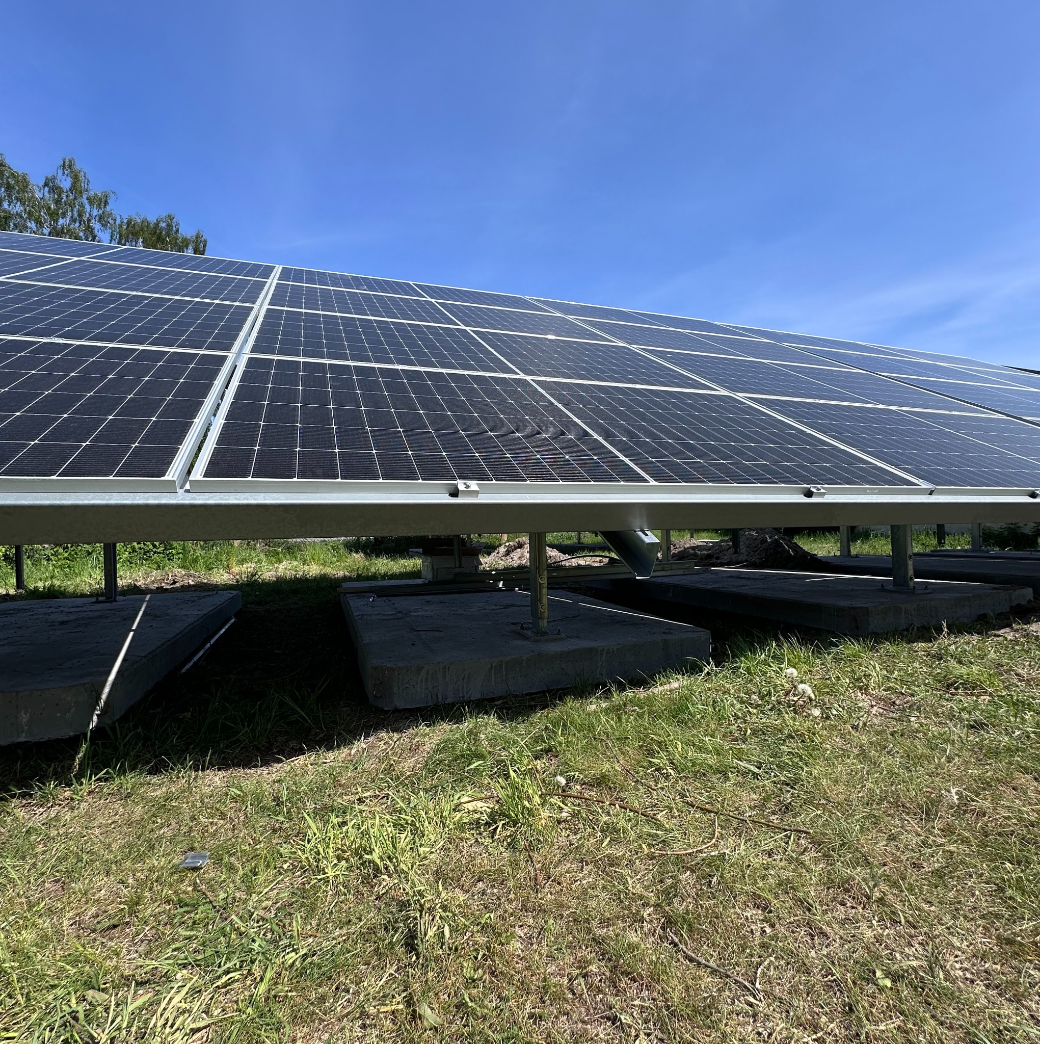 Rząd paneli słonecznych zamontowanych na betonowych fundamentach, widok z dołu na tle błękitnego nieba i zielonej trawy.