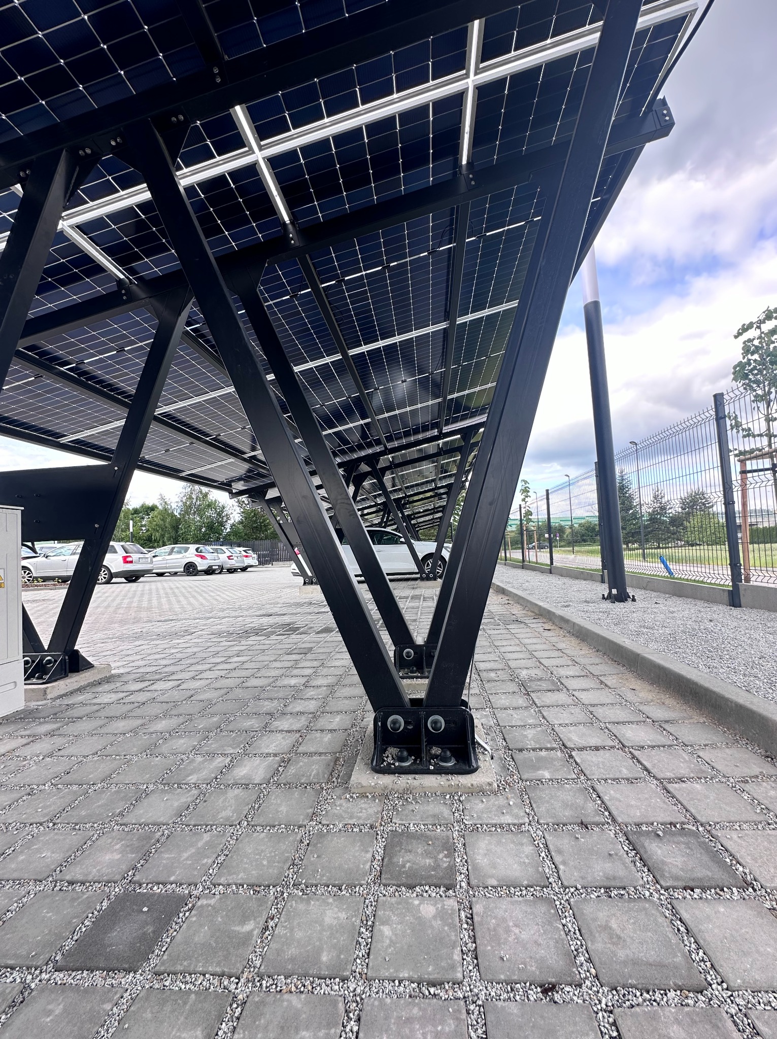 Zadaszenie parkingu wykonane z paneli fotowoltaicznych, widok z dołu na konstrukcję wspornikową z czarnych profili stalowych, szara kostka brukowa na parkingu, samochody w tle.