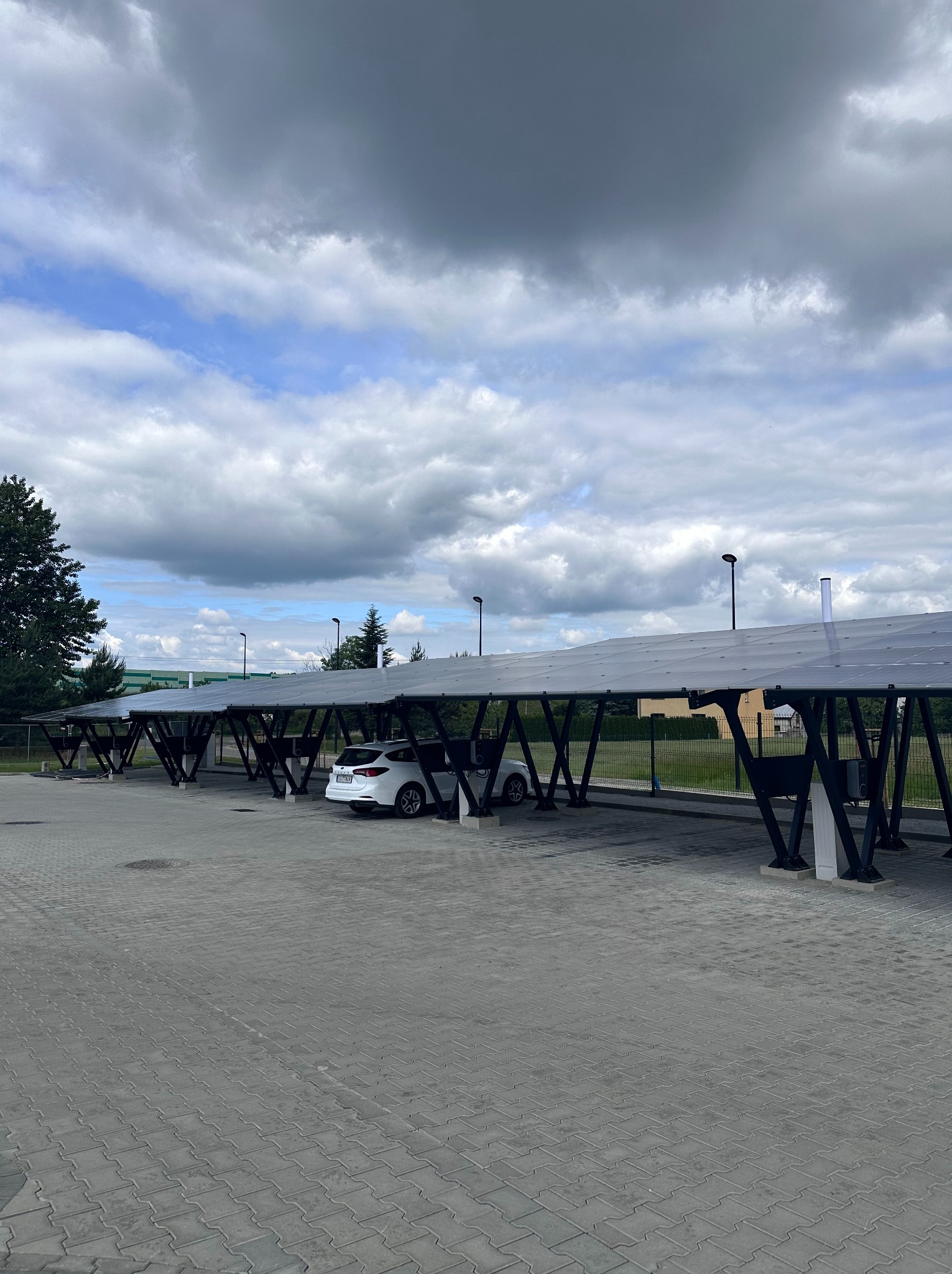 Zadaszony parking z panelami słonecznymi, biały samochód zaparkowany pod spodem, pochmurne niebo w tle.