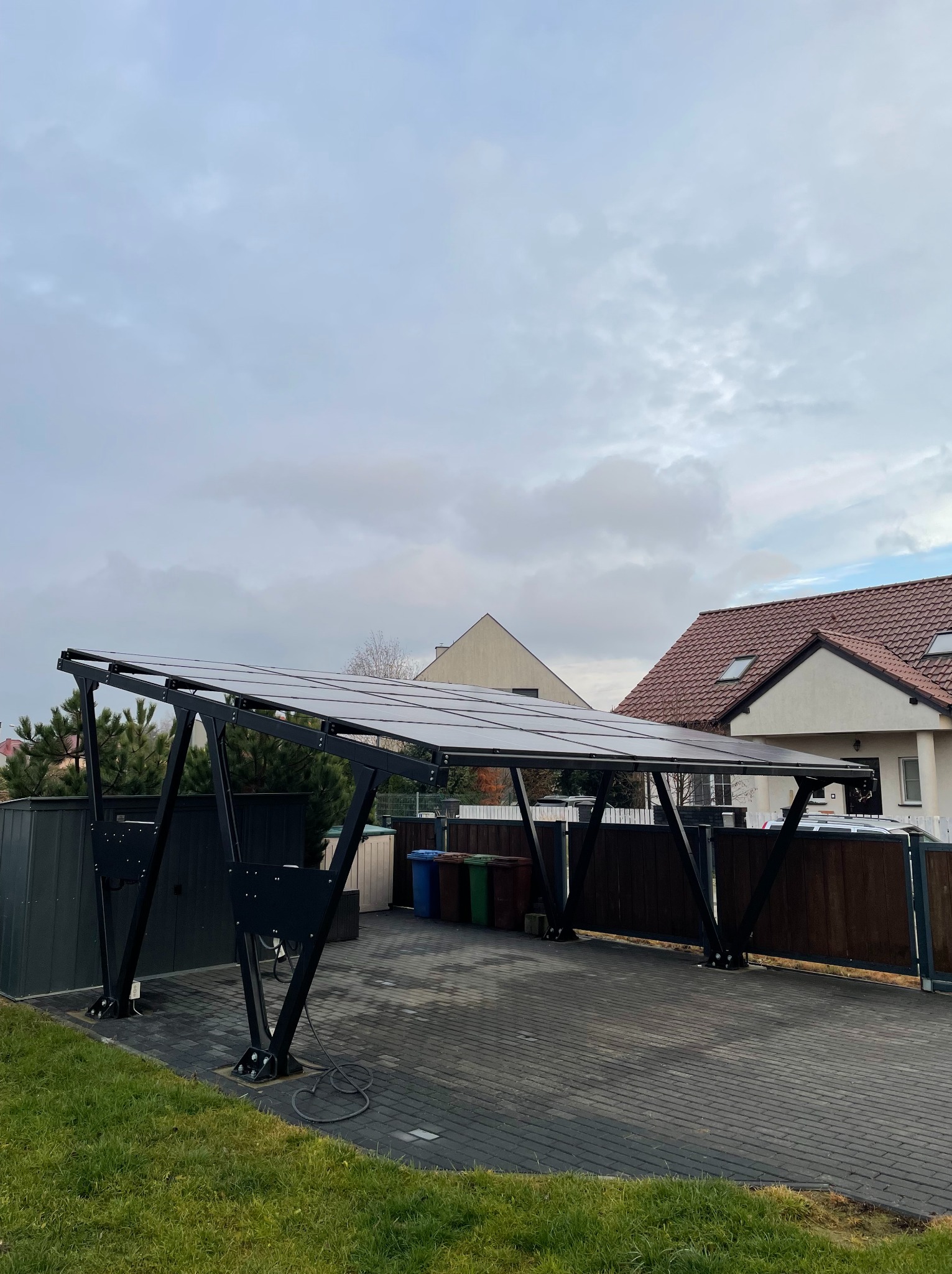 Zadaszenie z paneli słonecznych o czarnej konstrukcji na posesji z kostki brukowej, widoczne domy w tle.