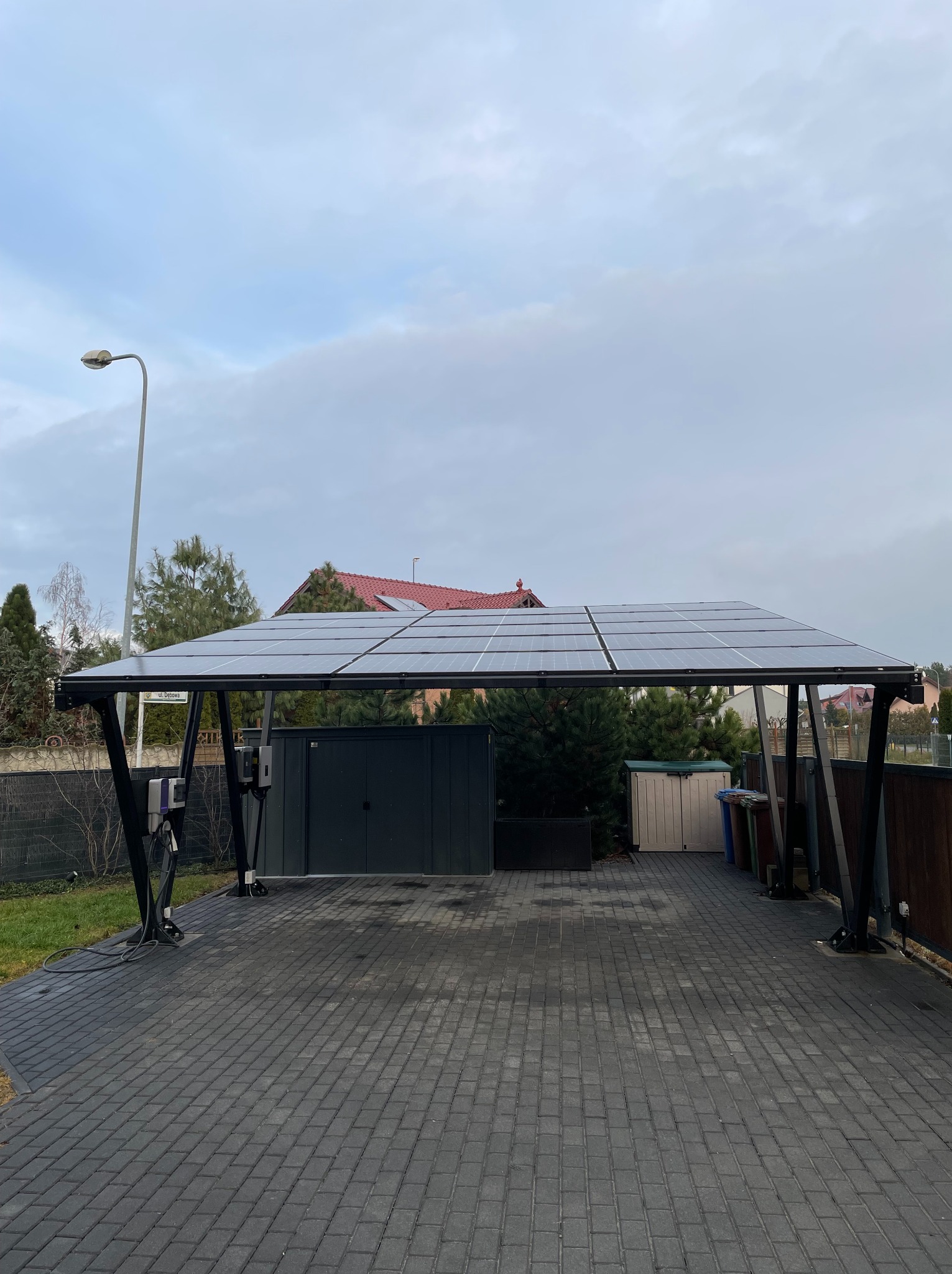 Zadaszenie z paneli słonecznych nad brukowanym podjazdem, widoczne stacje ładowania pojazdów elektrycznych, w tle dom z dachówką i drzewa, pochmurne niebo.