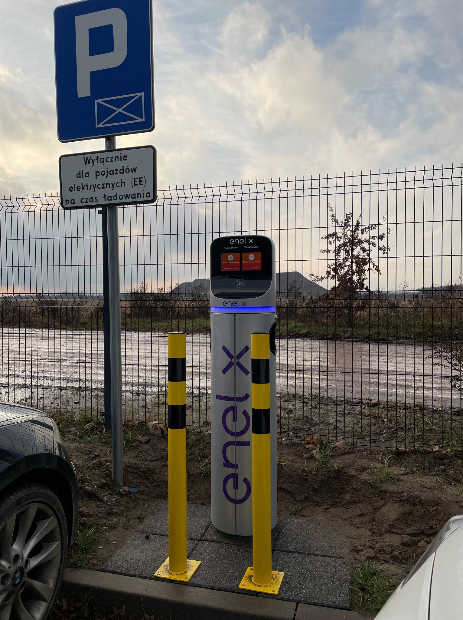 Stacja ładowania pojazdów elektrycznych Enel X z dwoma słupkami ochronnymi, umieszczona na parkingu z widocznym znakiem informacyjnym dla pojazdów elektrycznych.