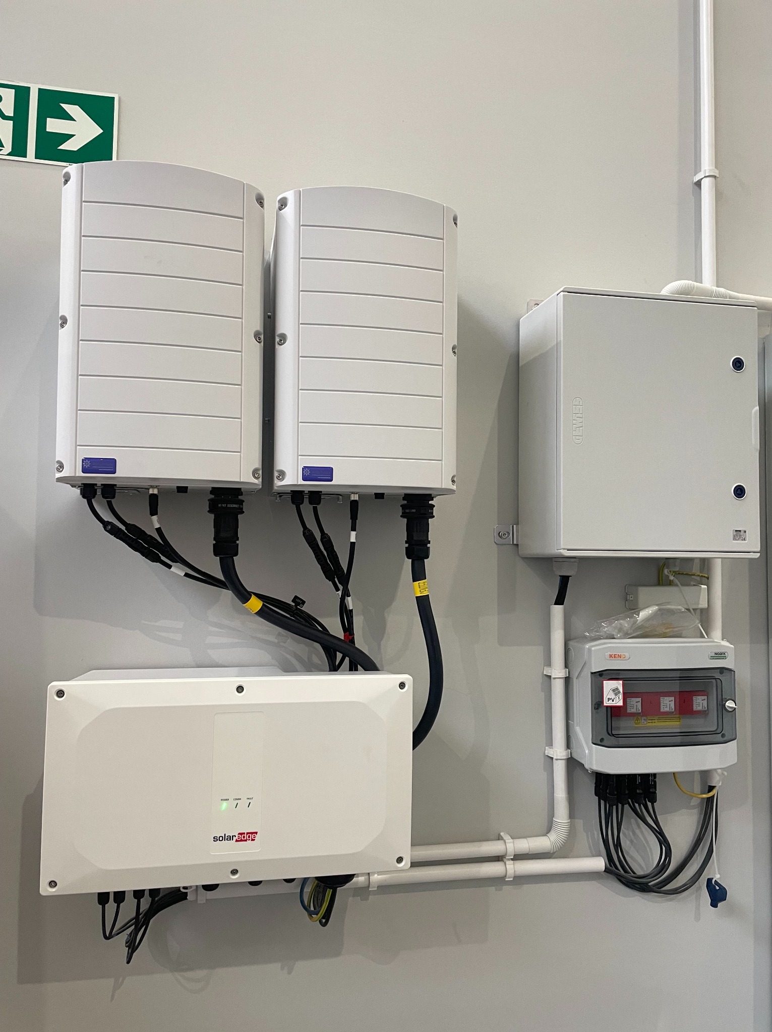 Dwa białe inwertery fotowoltaiczne SolarEdge z podłączonymi kablami, skrzynka przyłączeniowa i zabezpieczenia na szarej ścianie wewnątrz budynku, widoczny znak ewakuacyjny.