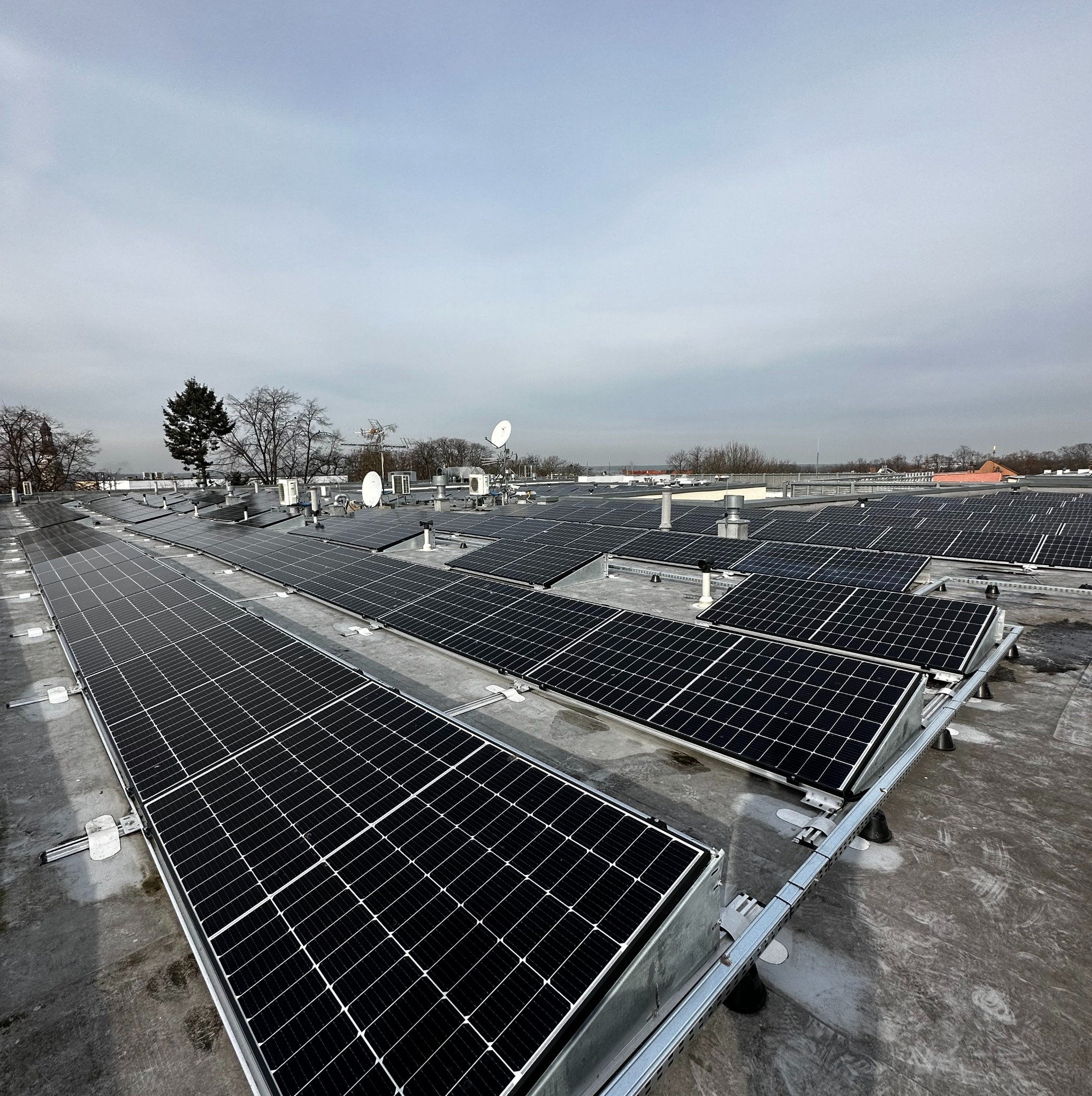 Rząd paneli słonecznych zamontowanych na płaskim dachu budynku przemysłowego, widoczne elementy mocujące i instalacje wentylacyjne, pochmurne niebo w tle.