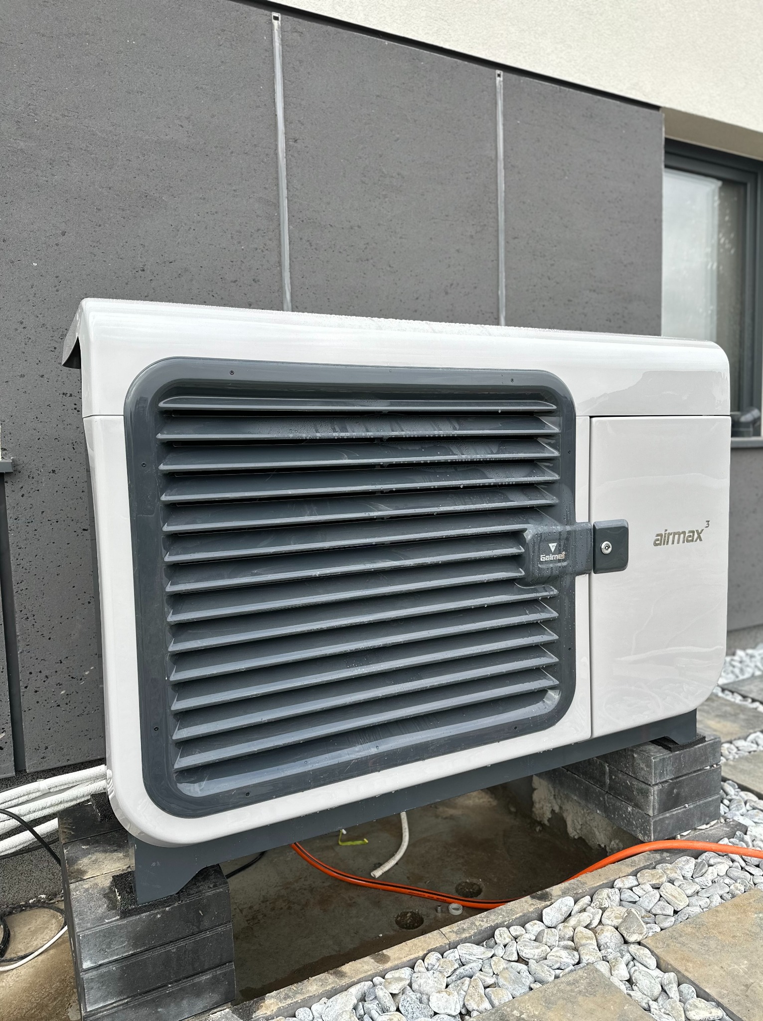 Biało-szara pompa ciepła Airmax 3 zamontowana na bloczkach betonowych przy elewacji budynku z widocznymi przewodami instalacyjnymi.