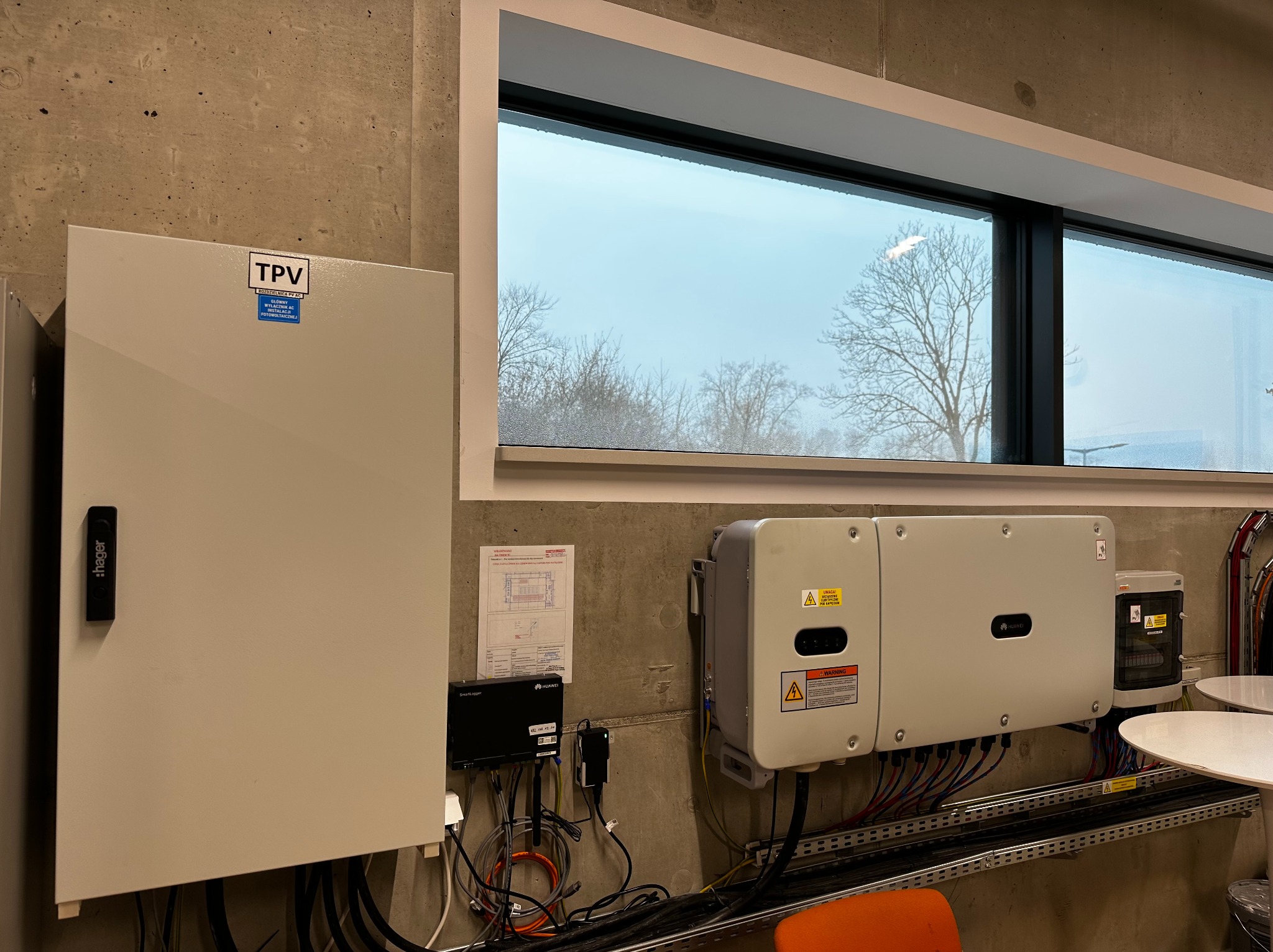 Rozdzielnica AC, inwerter i moduł komunikacyjny systemu fotowoltaicznego zamontowane na betonowej ścianie w pomieszczeniu z oknem, widoczne okablowanie i elementy montażowe.