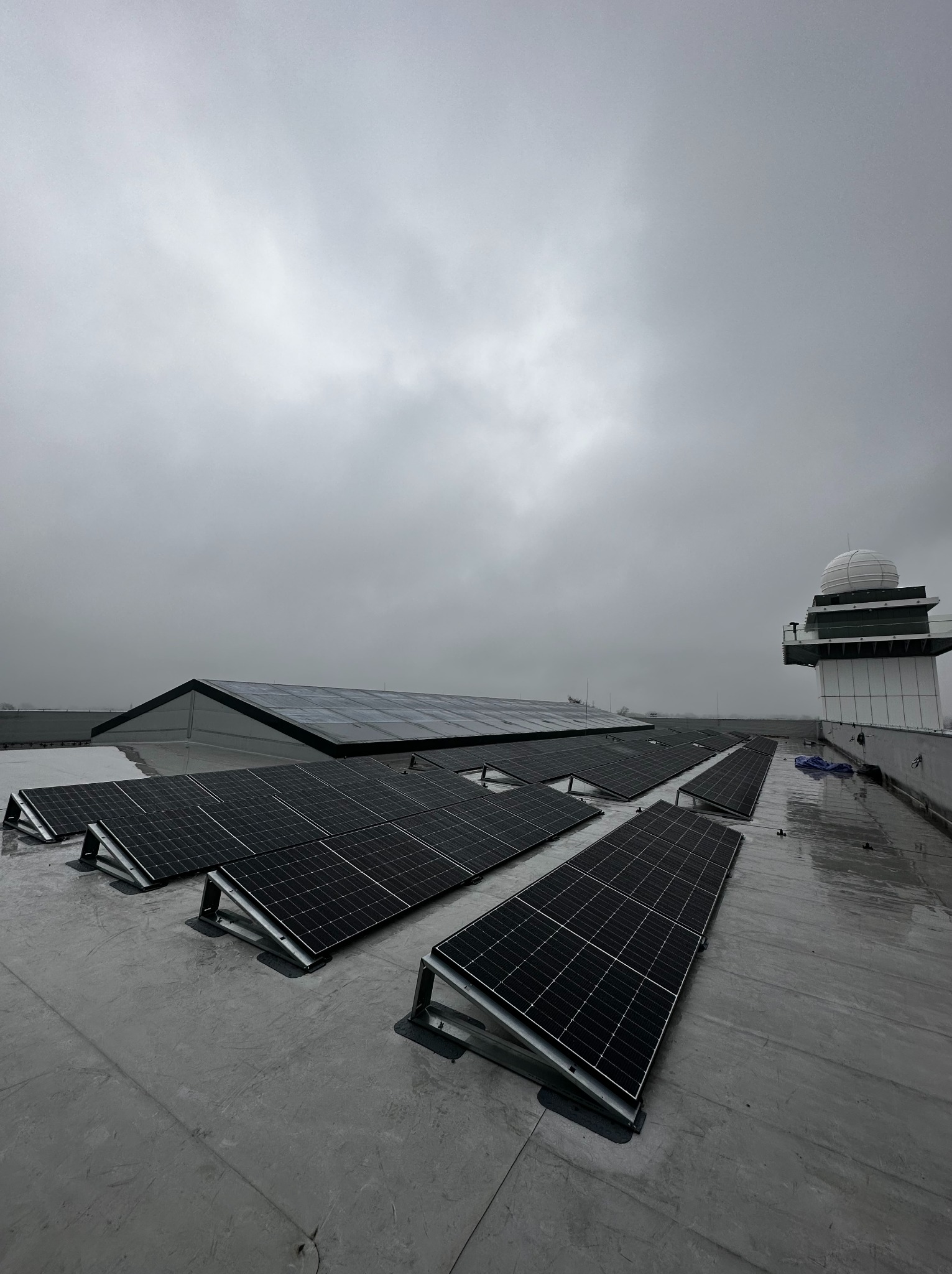 Montaż paneli fotowoltaicznych na płaskim dachu budynku z wieżą obserwacyjną w pochmurny dzień.