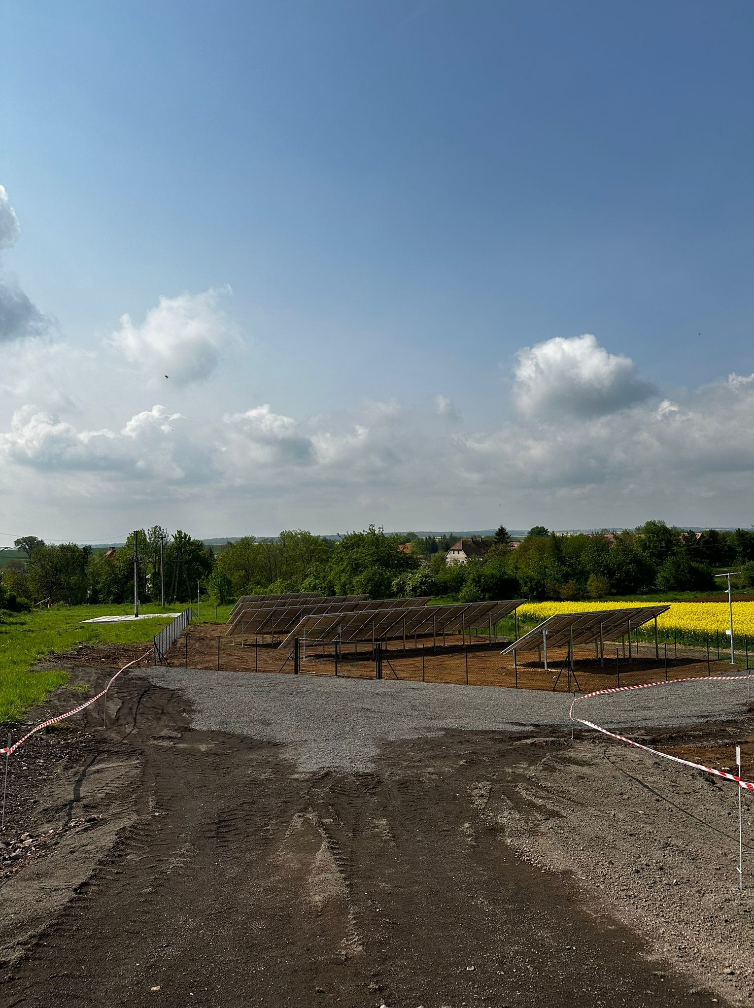 Montaż konstrukcji wsporczej paneli słonecznych na przygotowanym terenie, widok na pole rzepaku w tle, zabezpieczone taśmą ostrzegawczą.