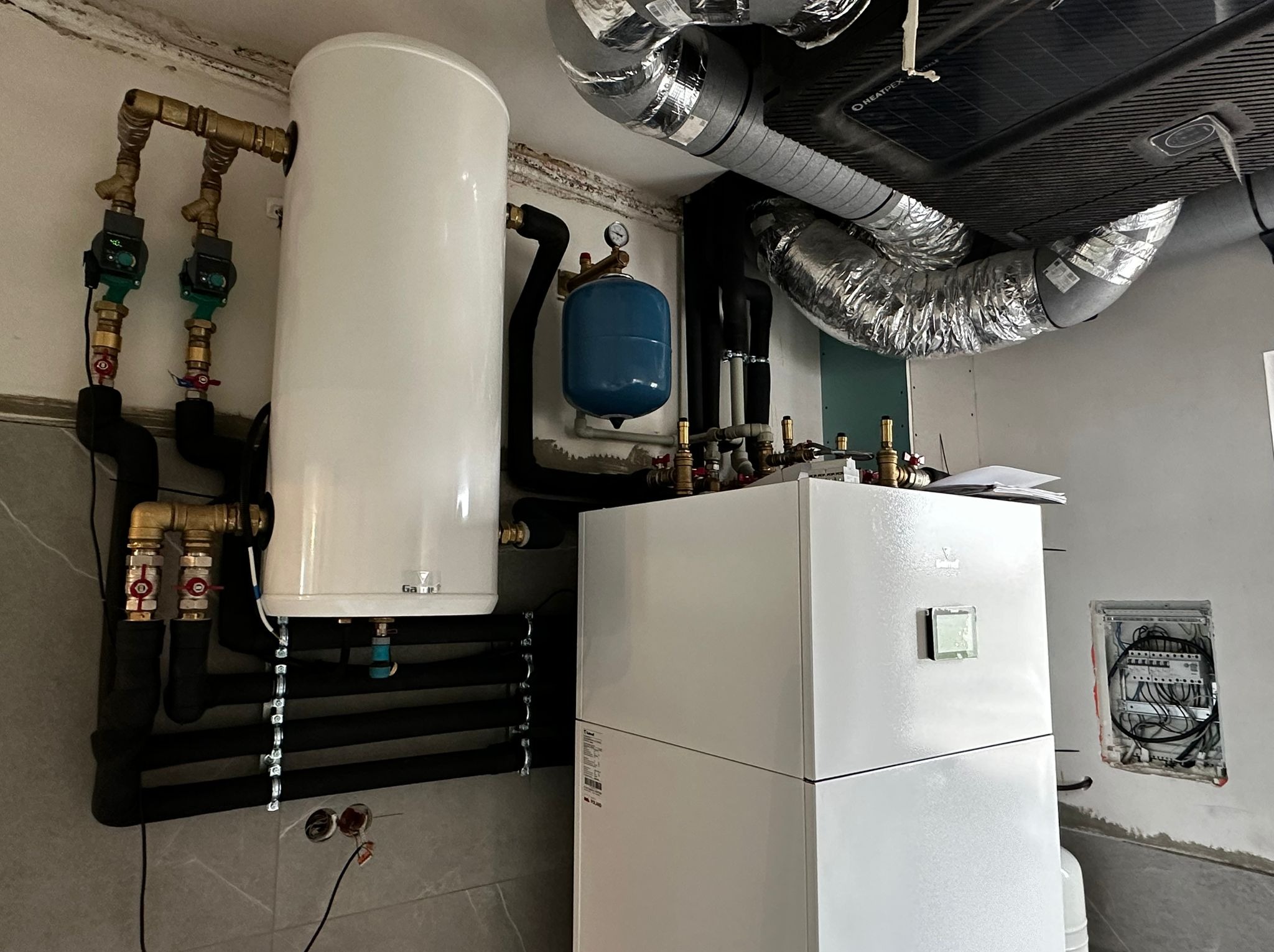 Biały zasobnik ciepłej wody i dwuczęściowa pompa ciepła w pomieszczeniu z widocznymi rurami, zaworami, manometrem i otwartą skrzynką elektryczną.