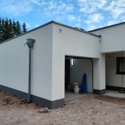 Dapir sp zoo - Jasna, otynkowana elewacja domu z garażem, widoczne rynny, kratki wentylacyjne, częściowo otwarta brama garażowa z widocznymi wewnątrz wiadrami z farbą i niebieską folią ochronną, kostka brukowa...