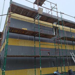 Dapir sp zoo - Dom w trakcie ocieplania, widoczne rusztowania, warstwy izolacji z wełny mineralnej i styropianu grafitowego ułożone naprzemiennie.