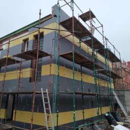 Dapir sp zoo - Budynek w trakcie ocieplania, widoczne rusztowania, warstwy izolacyjne w kolorach żółtym i grafitowym, nieotynkowana ściana z boku budynku, pochmurne niebo.