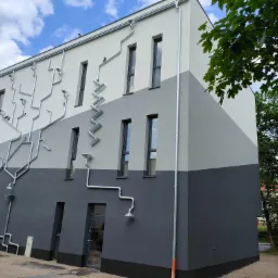 Dwukolorowa elewacja budynku z nietypowym systemem odprowadzania wody deszczowej w postaci dekoracyjnych rur i lejków.