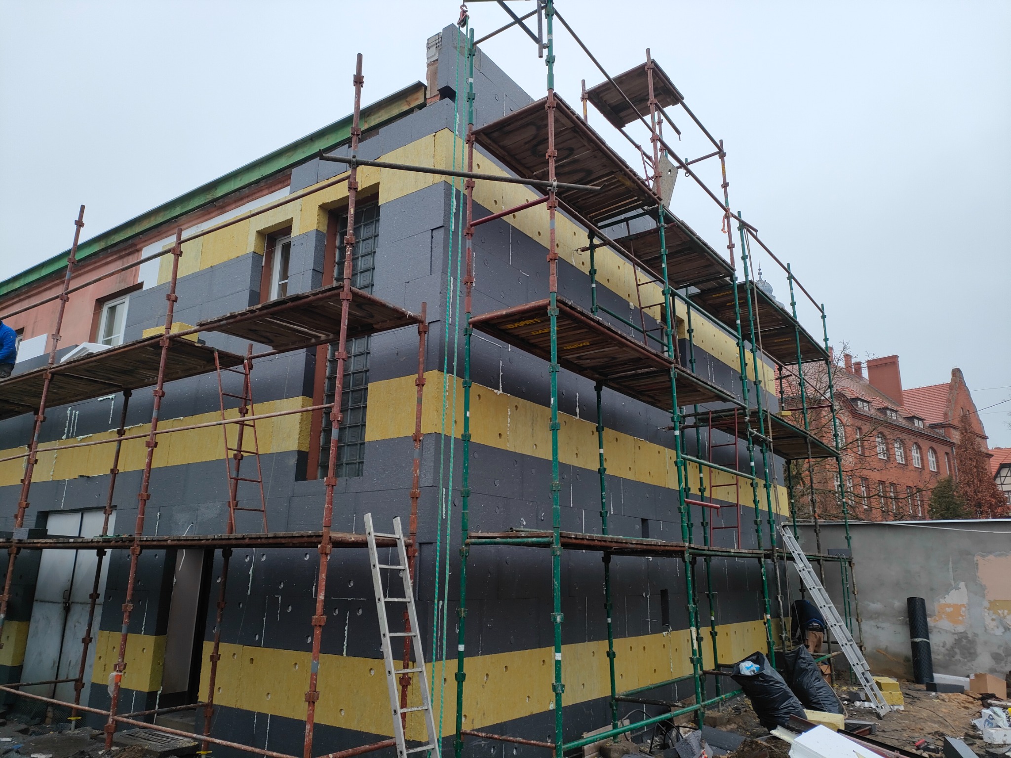 Budynek w trakcie ocieplania, widoczne rusztowania, warstwy izolacyjne w kolorach żółtym i grafitowym, nieotynkowana ściana z boku budynku, pochmurne niebo.